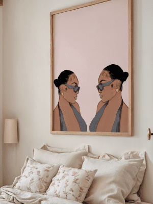 Framed mirrored portrait art print featuring two stylized profiles of women in sunglasses and a halter top, displayed above a bed.