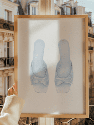 Framed watercolor art print featuring a pair of elegant light blue satin slip-on mule heels with bows, held in front of a sunny window overlooking a Parisian apartment building balcony.