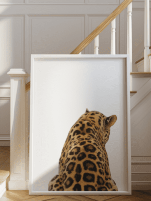 A close-up photo of the back and upper body of a leopard, showcasing its spotted fur, printed in a white frame leaning on a light wood floor near a white staircase.