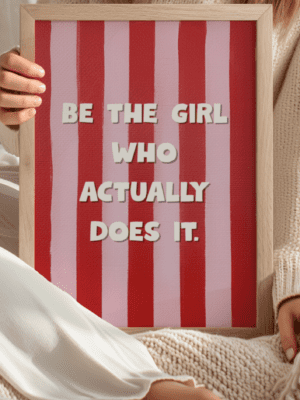 A motivational wall art print in a light wood frame featuring the phrase "BE THE GIRL WHO ACTUALLY DOES IT." The background is a bold, vertical pattern of alternating pink and deep red stripes. The print is being held by a woman wearing a cream-colored sweater, resting on a white blanket or cloth.