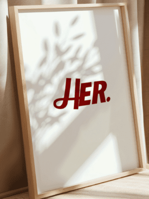 Minimalist wall art print featuring the word "HER." in a bold red stylized font on a clean white background, framed in a light wood frame.