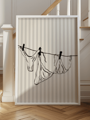 A vertical, framed, black line drawing of three pieces of delicate underwear hanging by clothespins on a tight clothesline. The artwork is set against a soft, neutral-striped background and is displayed propped up near a wooden staircase with white trim.