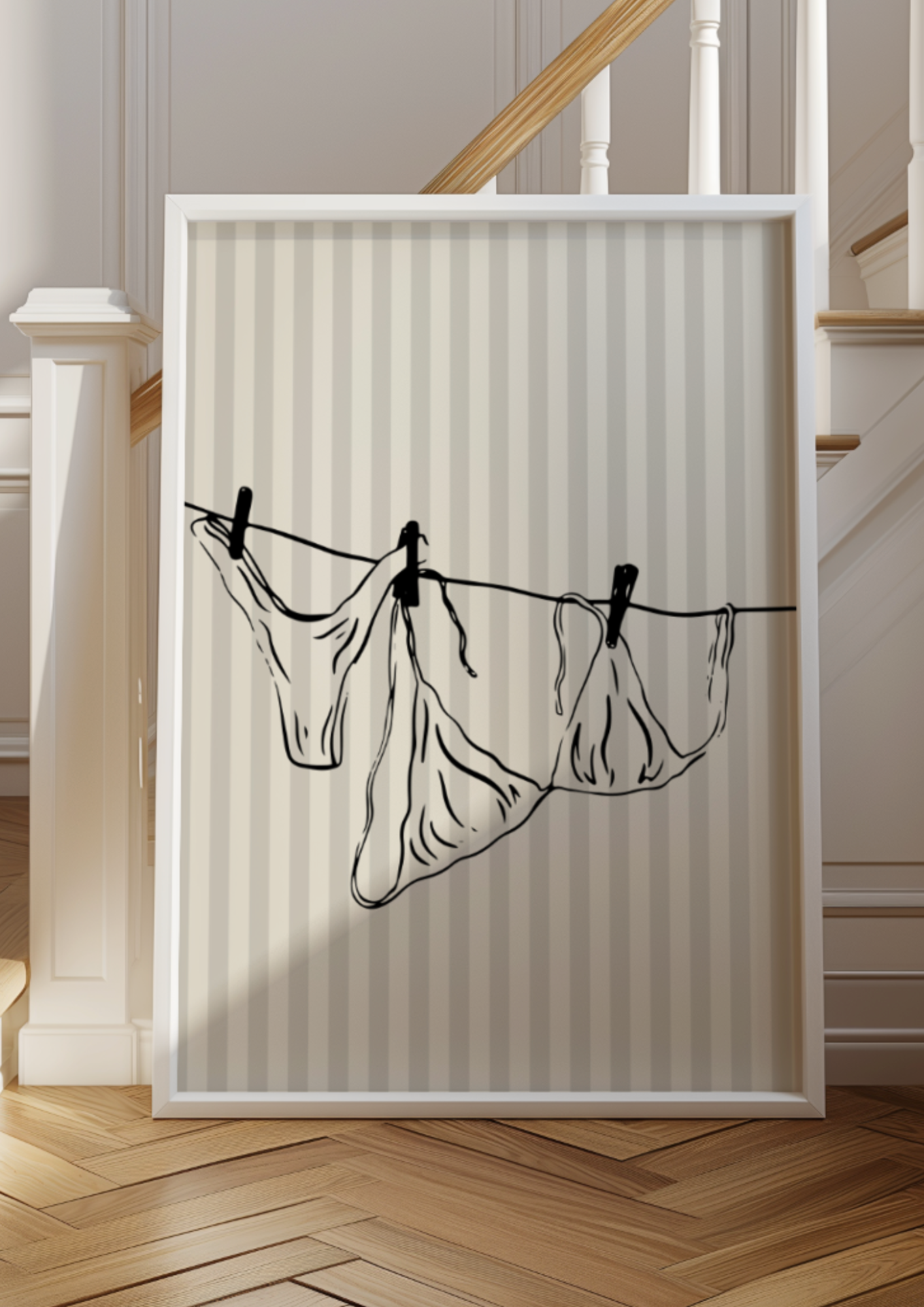 A vertical, framed, black line drawing of three pieces of delicate underwear hanging by clothespins on a tight clothesline. The artwork is set against a soft, neutral-striped background and is displayed propped up near a wooden staircase with white trim.