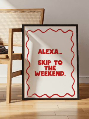 A black-framed art print leaning against a light wood chair. The print has a white background and features the phrase "ALEXA... SKIP TO THE WEEKEND." in bold, red, block letters, surrounded by a wavy red border. The image is taken indoors on a wooden floor, showing bright natural light.