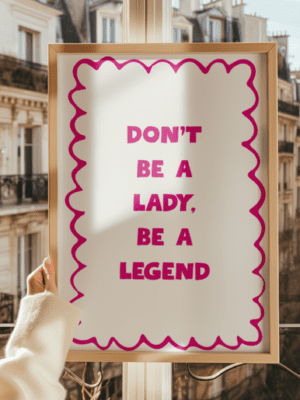 A close-up photograph of a framed print featuring the motivational quote "DON'T BE A LADY, BE A LEGEND" in bold pink text with a decorative scalloped pink border, held up against a sunny window overlooking Parisian buildings.