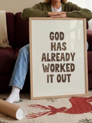 Woman leaning on couch holding large framed poster with "God Has Already Worked It Out" text in leopard print pattern, next to rolled poster tube on reindeer-patterned rug in cozy home setting.