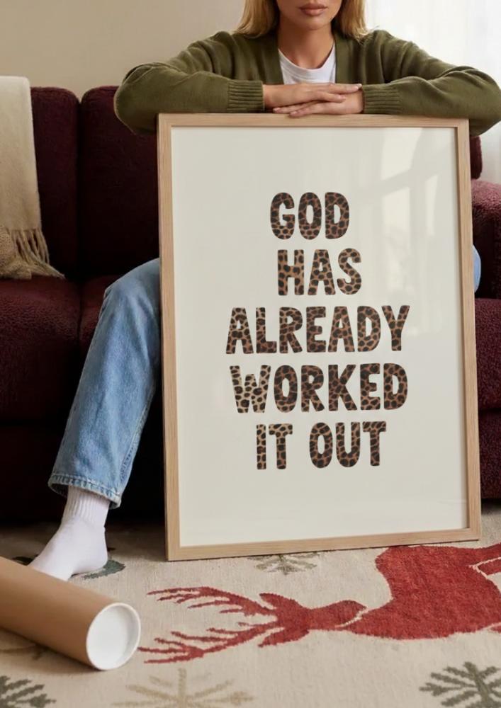 Woman leaning on couch holding large framed poster with "God Has Already Worked It Out" text in leopard print pattern, next to rolled poster tube on reindeer-patterned rug in cozy home setting.